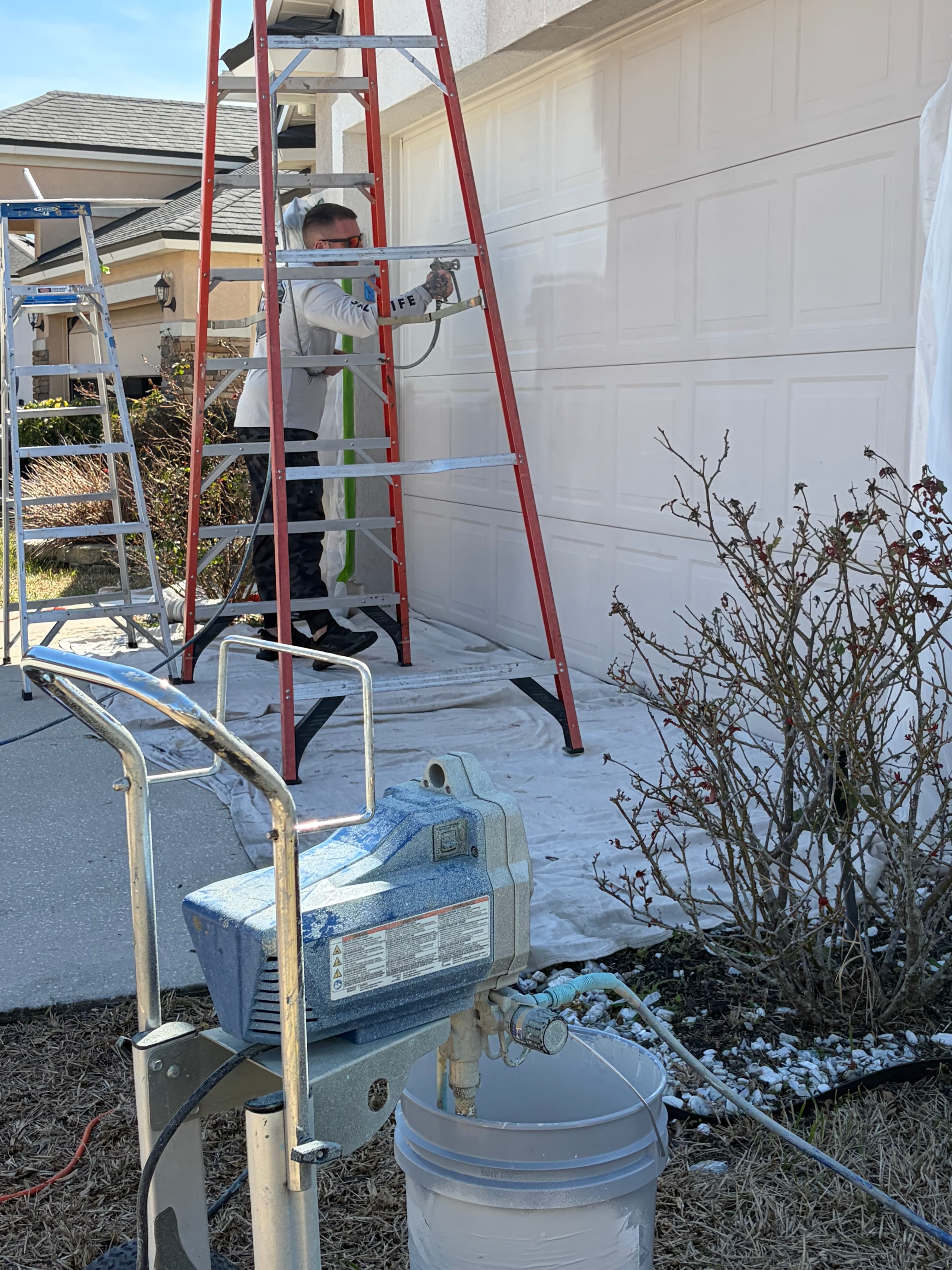 Man on a ladder using a paint sprayer to paint a white garage door.