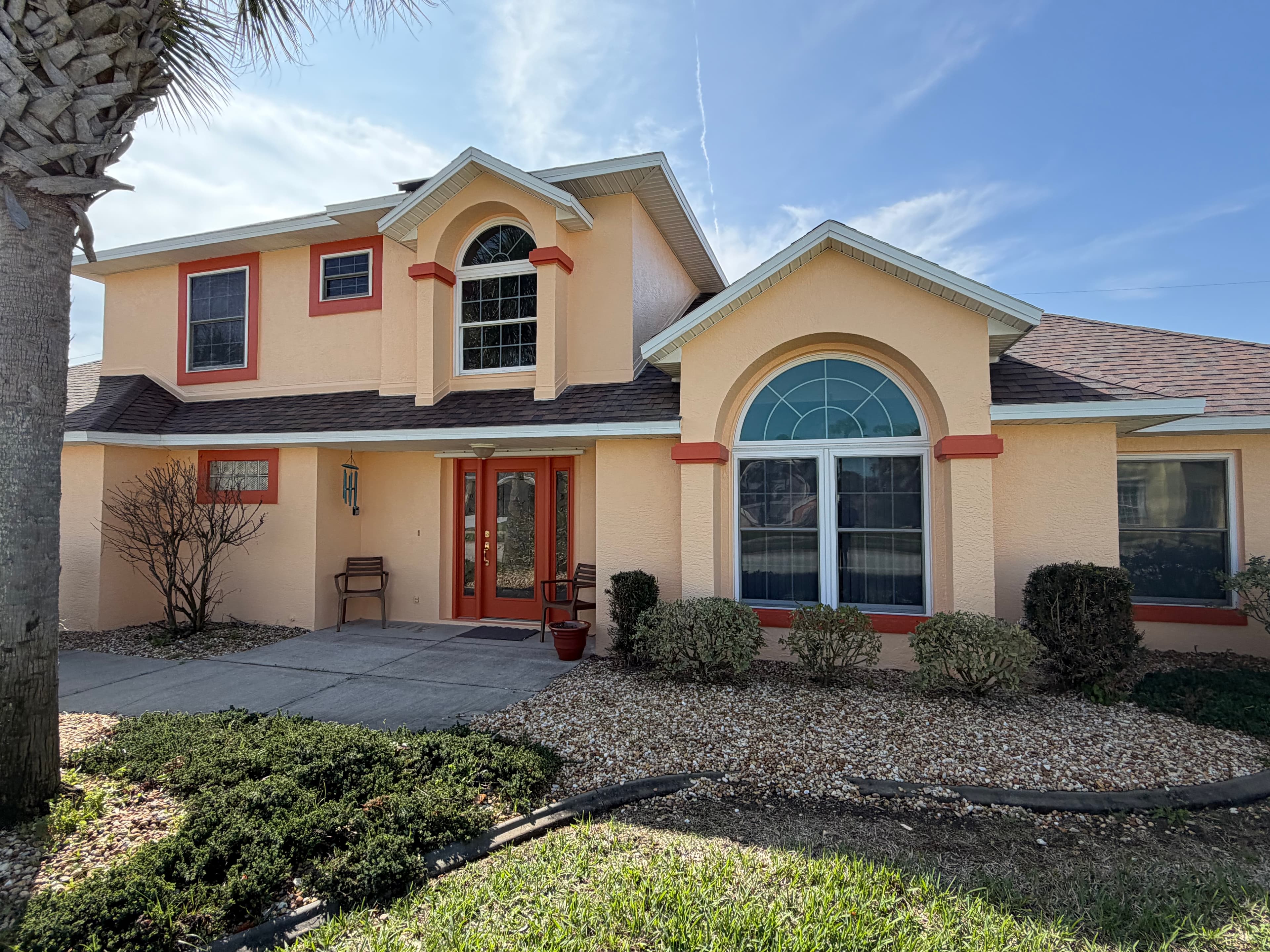 Two-story peach house with red trim, large arched windows, and a palm tree in front.