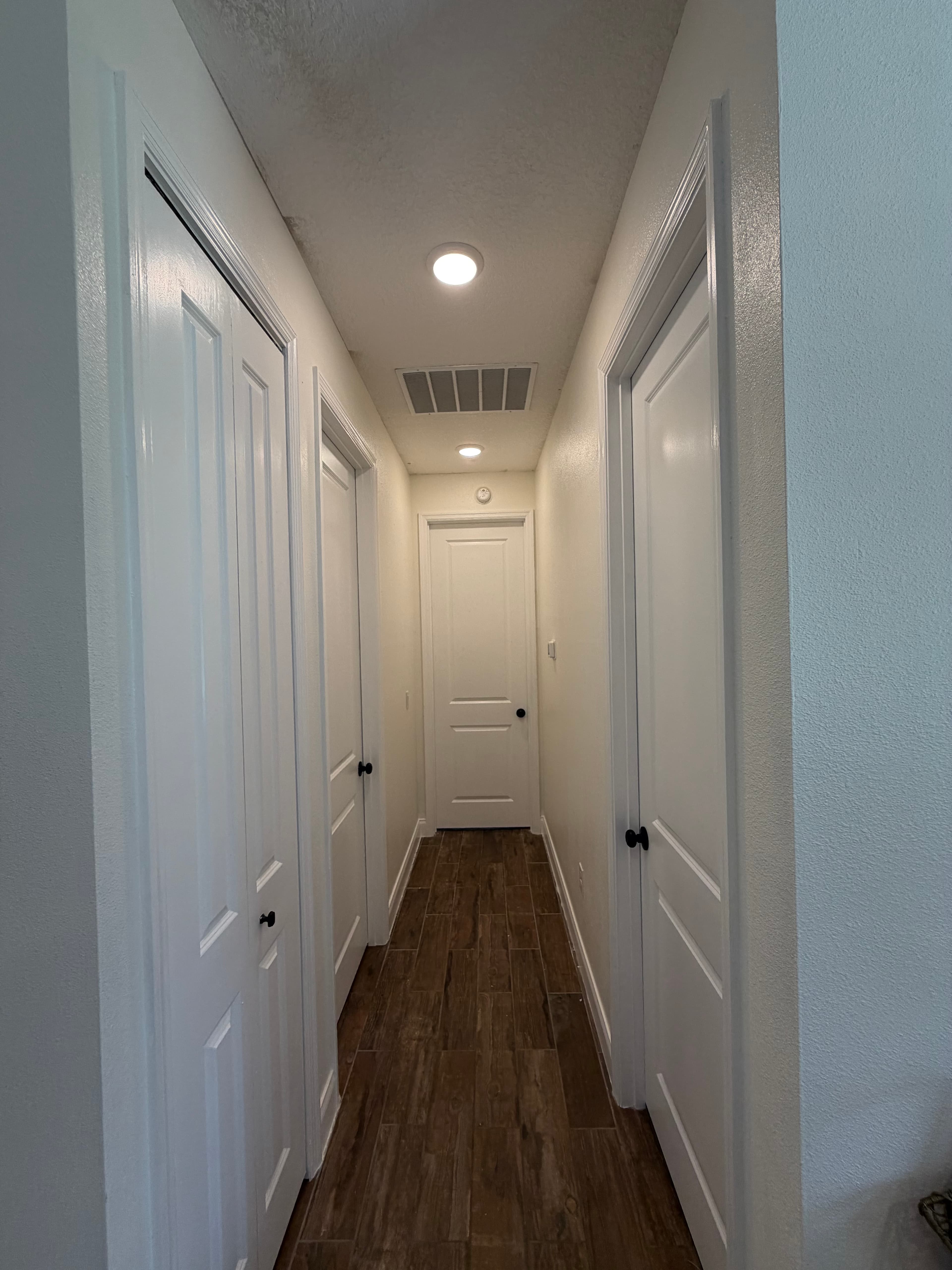 Narrow white hallway featuring dark wood-textured tile floors and several closed white doors.