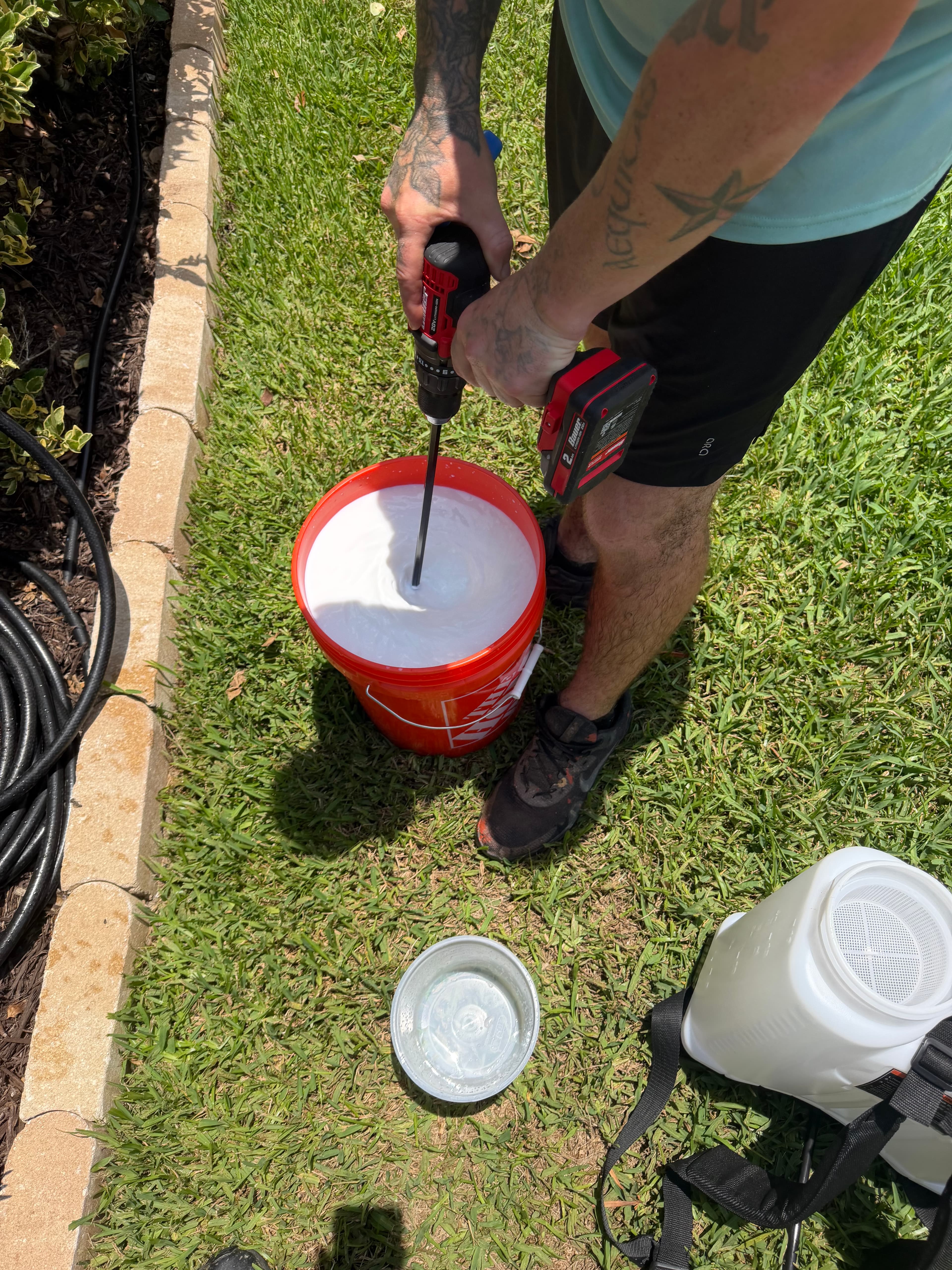Tattooed person using a power drill to mix white liquid in a bucket on grass.
