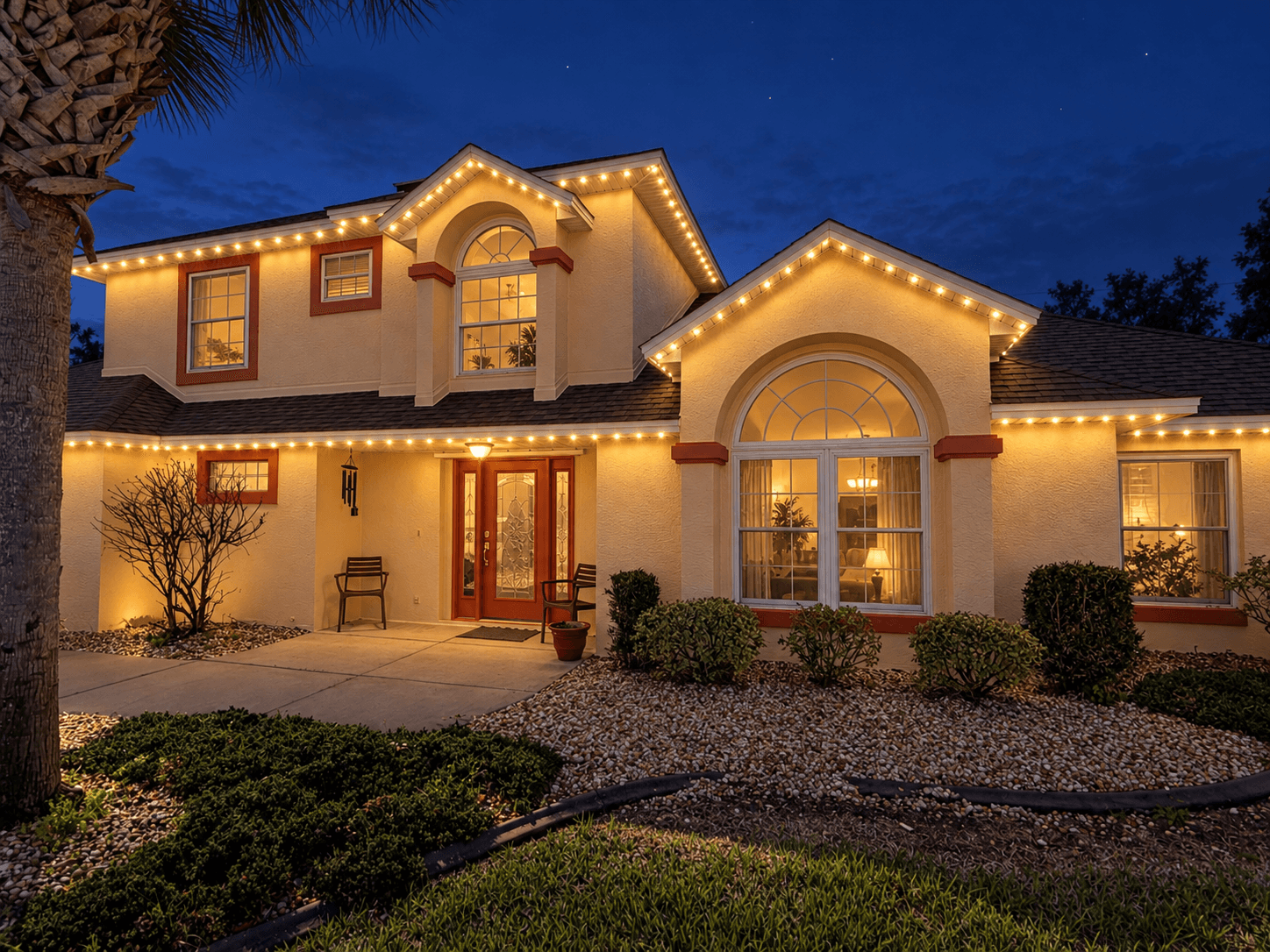 Two-story beige house illuminated by warm white string lights along the roofline at twilight.